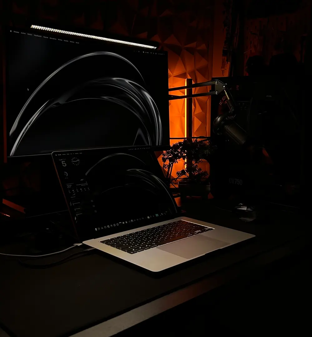Mi mesa de trabajo: un monitor con la web de Kalani Travels abierta y un portátil con código, iluminados por una luz naranja